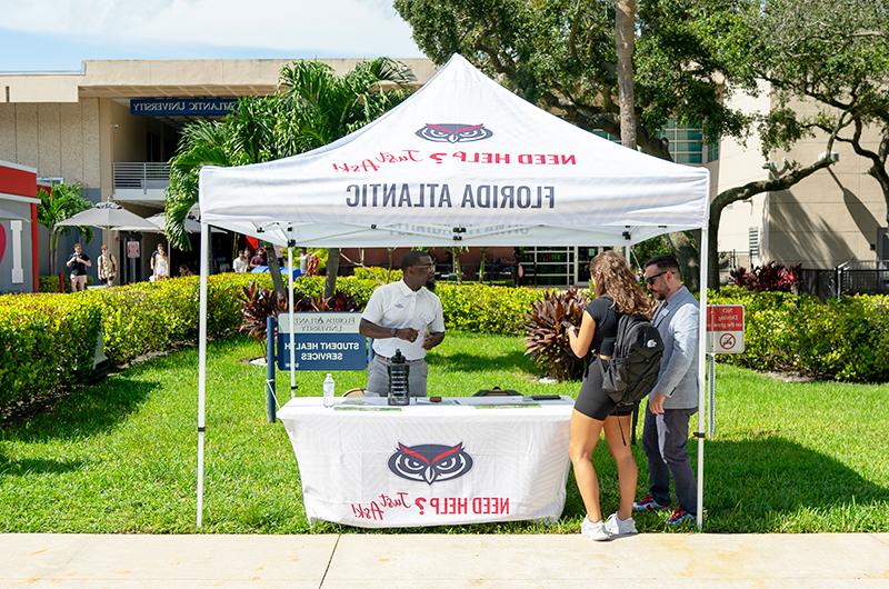 a white tent with FAU logos on it placed on the grass next to a busy walkway on campus, with staff members helping students find their way